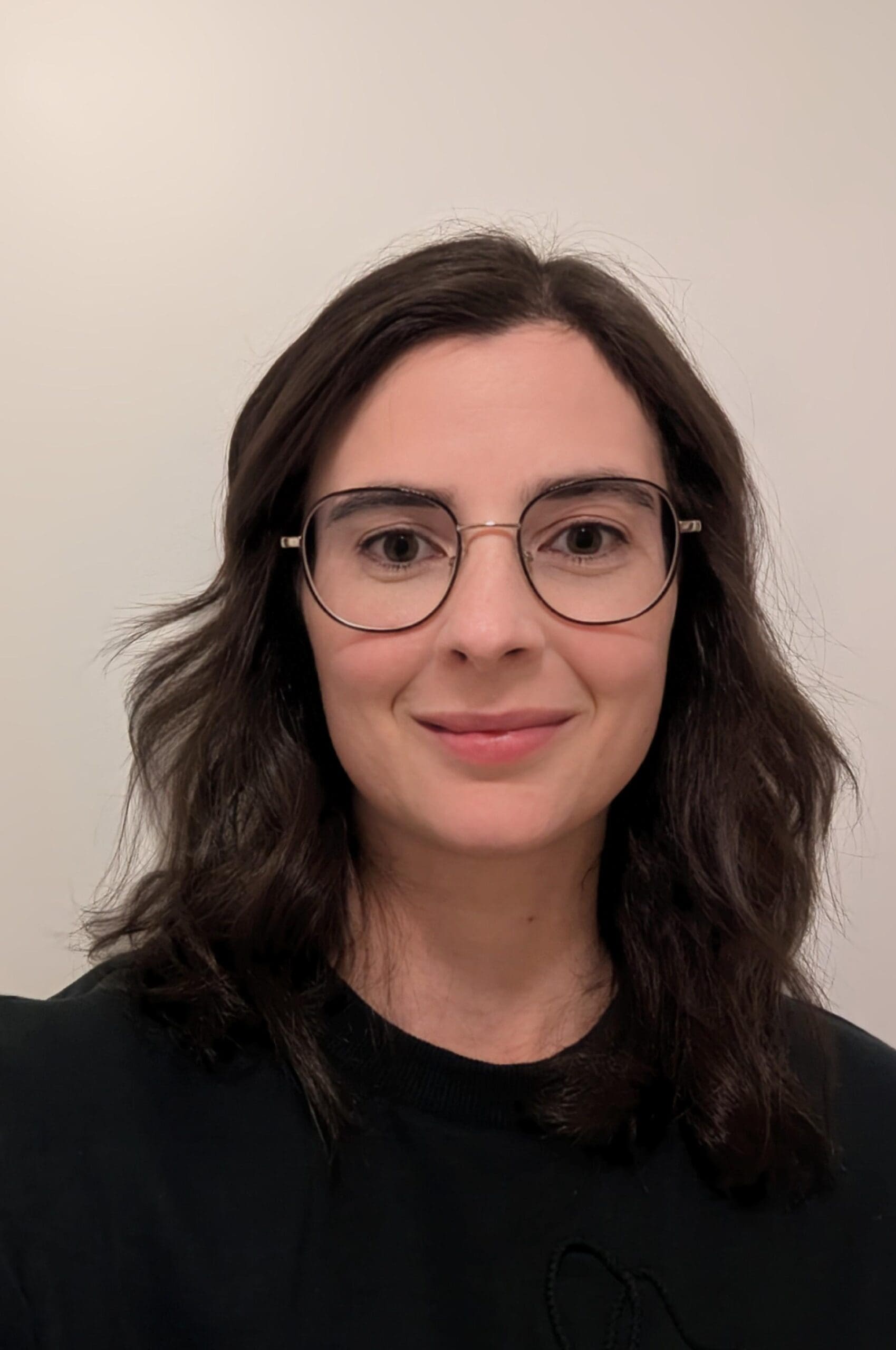Une femme aux cheveux noirs ondulés et aux lunettes sourit légèrement à l'appareil photo. Elle porte un haut noir et se tient devant un fond uni de couleur claire.
