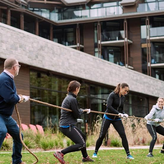 Quatre personnes, trois femmes et un homme, jouent au tir à la corde à l'extérieur, sur une pelouse située devant un immeuble moderne. Toutes sont vêtues de tenues de sport et semblent prendre plaisir à cette activité.