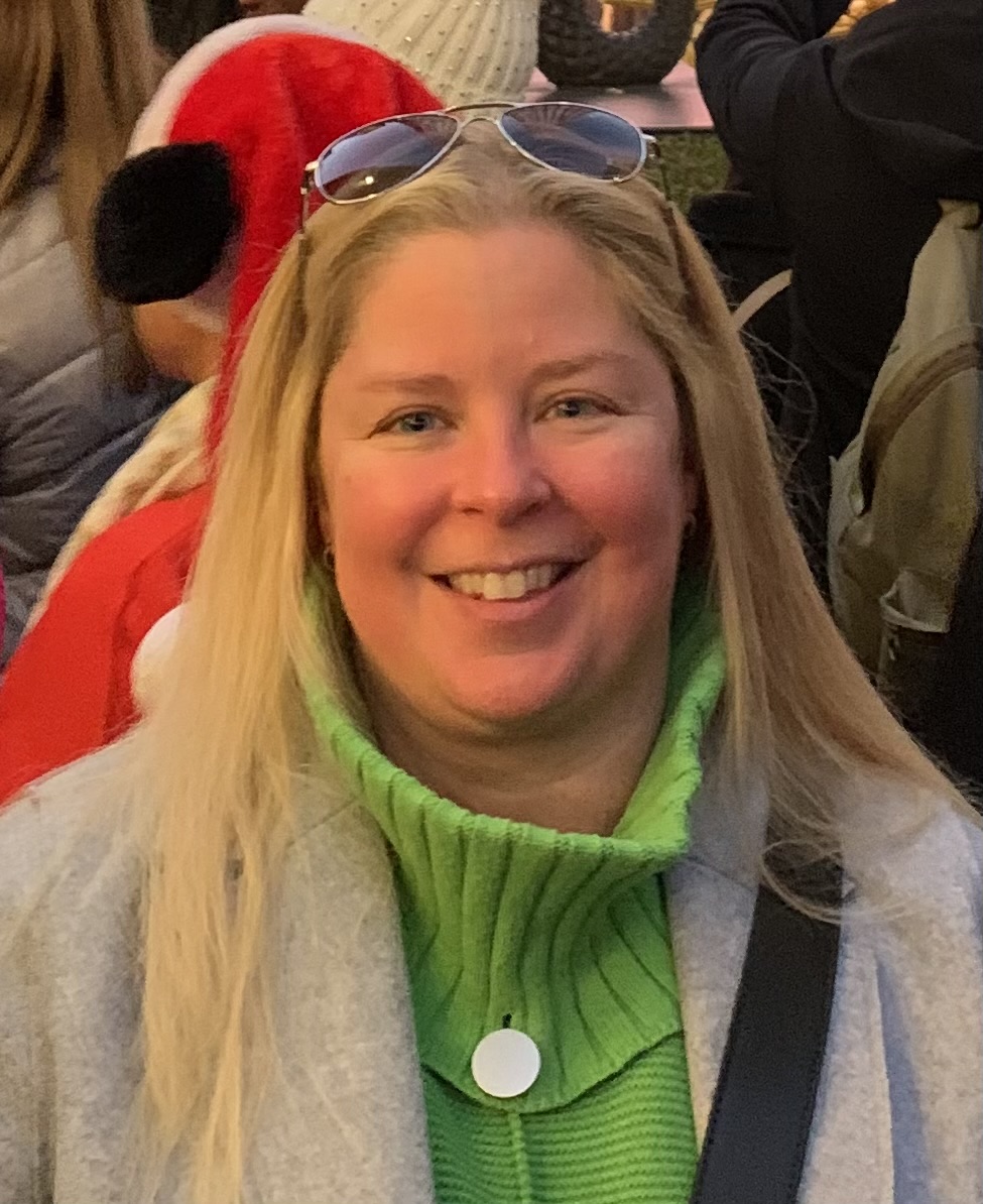 Une femme souriante aux longs cheveux blonds porte un pull à col roulé vert, un manteau gris et des lunettes de soleil posées sur la tête. Elle se tient dehors au milieu d'une foule, avec derrière elle une personne déguisée en Père Noël.