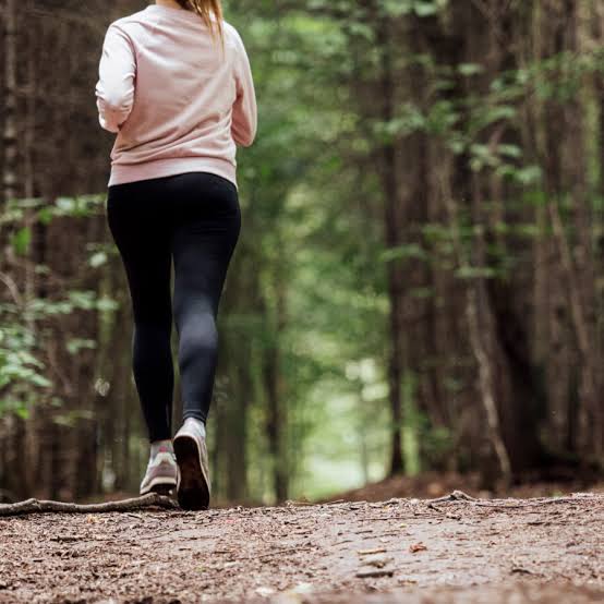 Une personne vêtue d'un sweat-shirt rose pâle et d'un legging noir fait son jogging sur un sentier forestier, au milieu de grands arbres et de la verdure. L'atmosphère est paisible et le chemin est recouvert de terre et de petites branches.